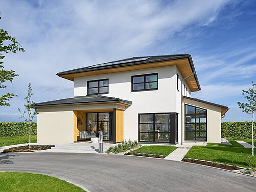 Ein modernes, zweistöckiges Haus mit einem flachen Dach und großen Fenstern. Die Fassade ist überwiegend weiß mit Holzdetails. Im Vordergrund befinden sich ein gepflasterter Weg und ein Rasen, umgeben von kleineren Bäumen und Hecken. Der Himmel ist klar und blau.